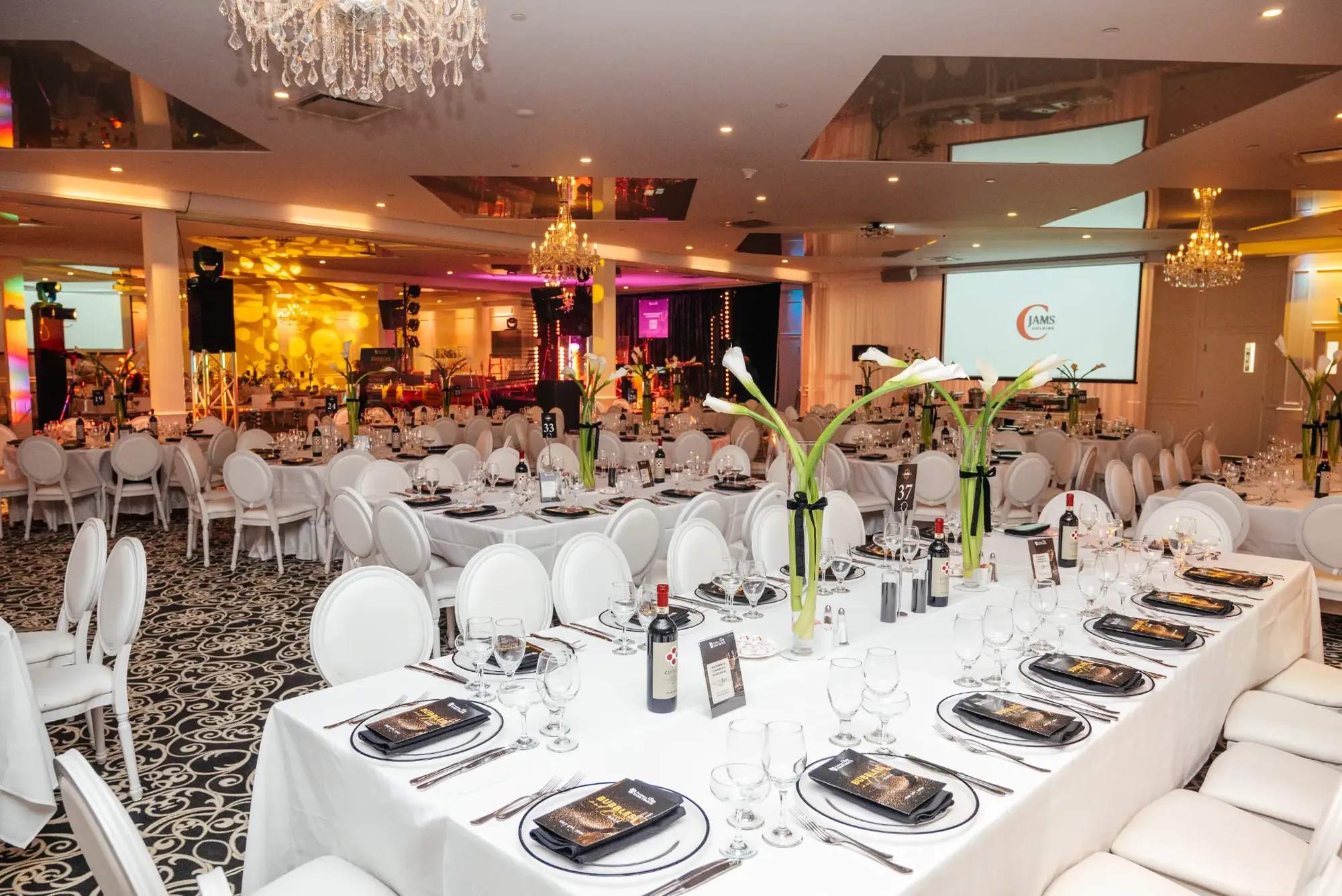 Grande salle de réception aménagée avec tables blanches et hauts centres de table floraux.