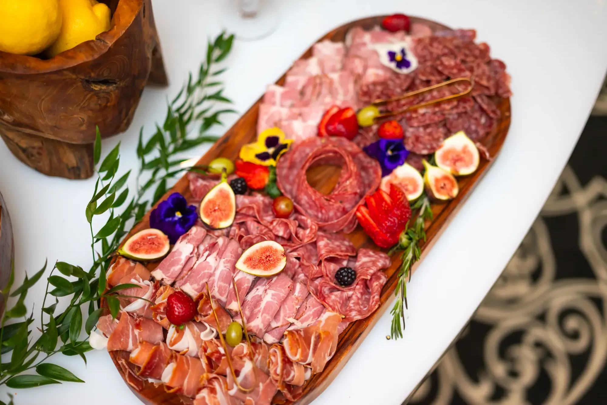 Longue planche de charcuterie garnie de fruits frais et de fleurs comestibles.